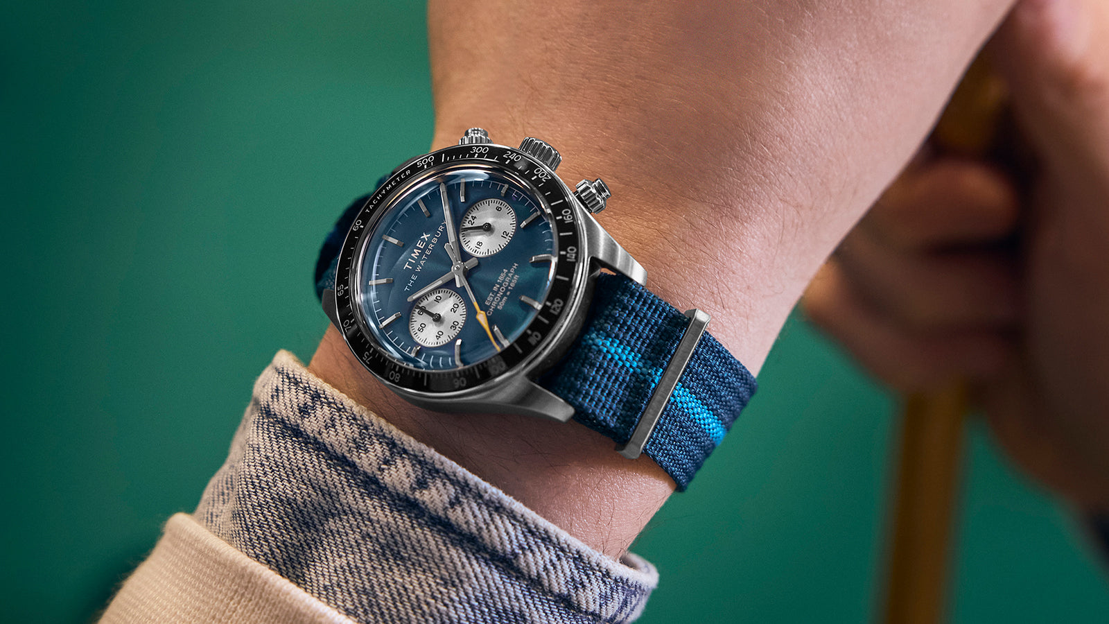 Timex Waterbury Heritage Chronograph with blue dial and striped blue strap worn on wrist, styled with a denim jacket sleeve.