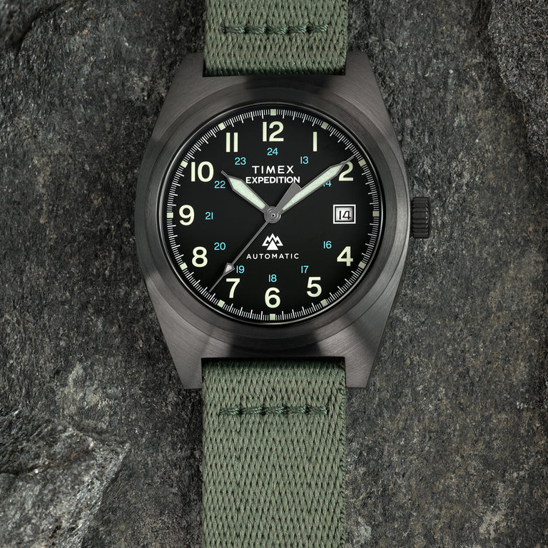 Timex Expedition automatic field watch with a black dial, luminous Arabic numerals, date window, brushed dark case, and green fabric strap, photographed on a stone surface.