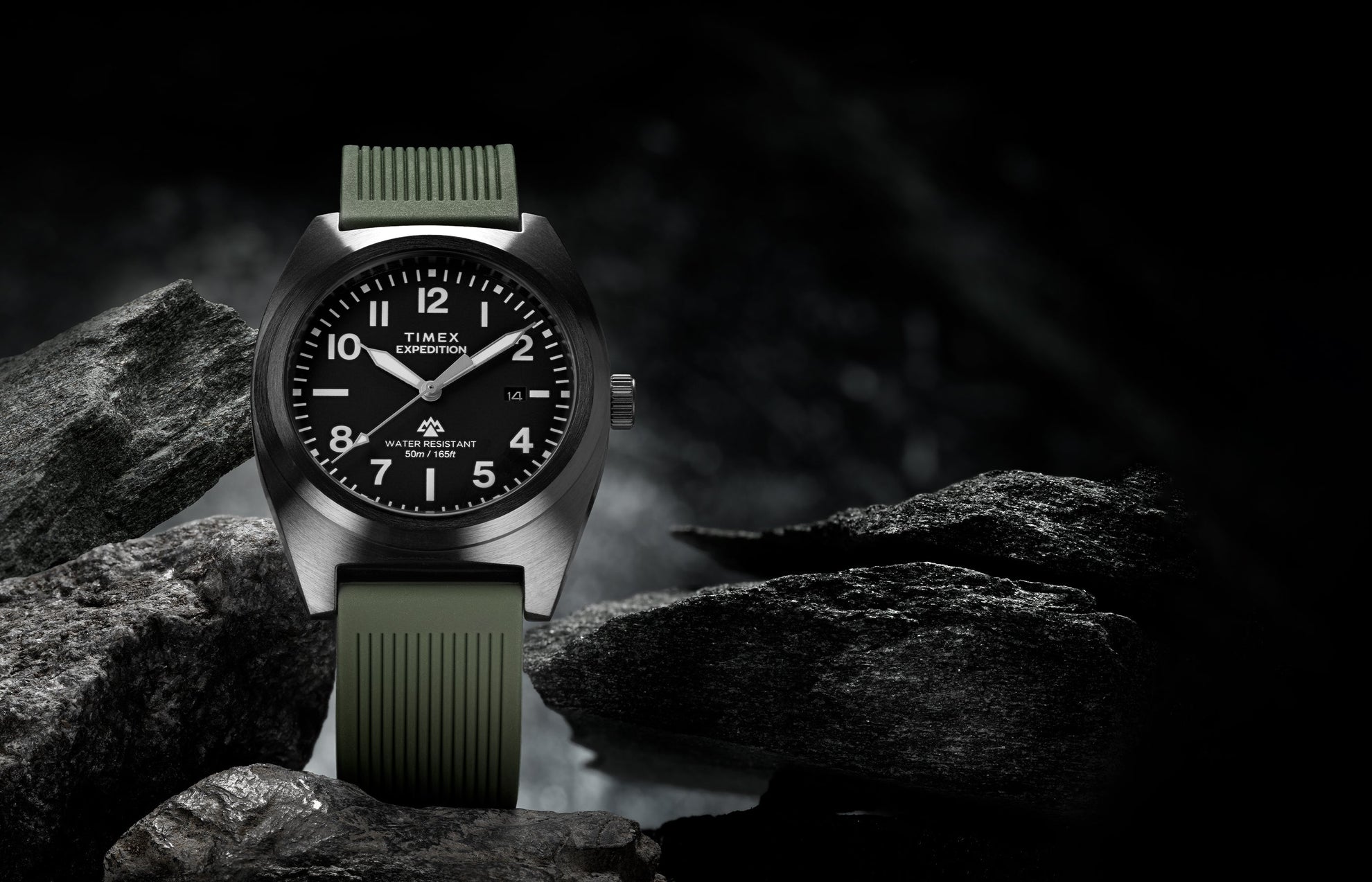 Timex Expedition® Capstone watch with a black dial and olive green silicone strap, displayed among rugged dark rocks. The watch face shows bold white numerals and hands, highlighting its adventure-ready design and 50-meter water resistance.
