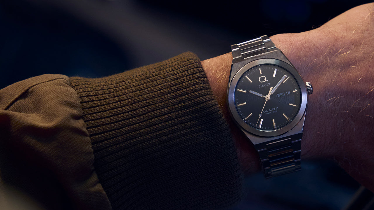 Wrist shot of the Q Timex® Continental Day and Date worn under a brown jacket sleeve. The stainless-steel bracelet and dark dial pair effortlessly for a timeless, understated style suited for both casual and formal wear.