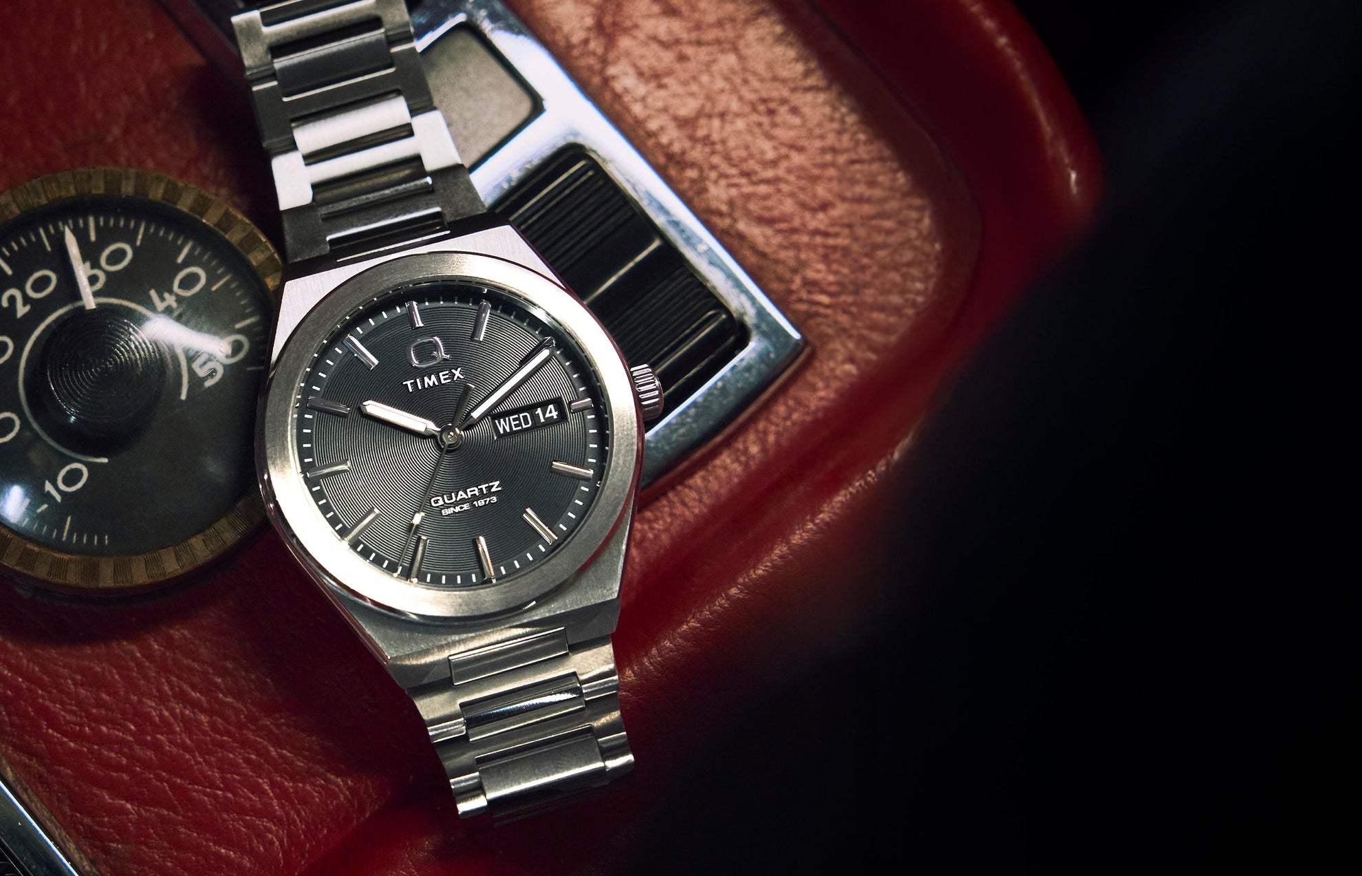 A Q Timex® Continental Day and Date watch with a brushed stainless-steel case and bracelet rests on a red vintage car dashboard. The dark dial displays the day and date window at 3 o’clock, blending retro automotive style with modern sophistication.
