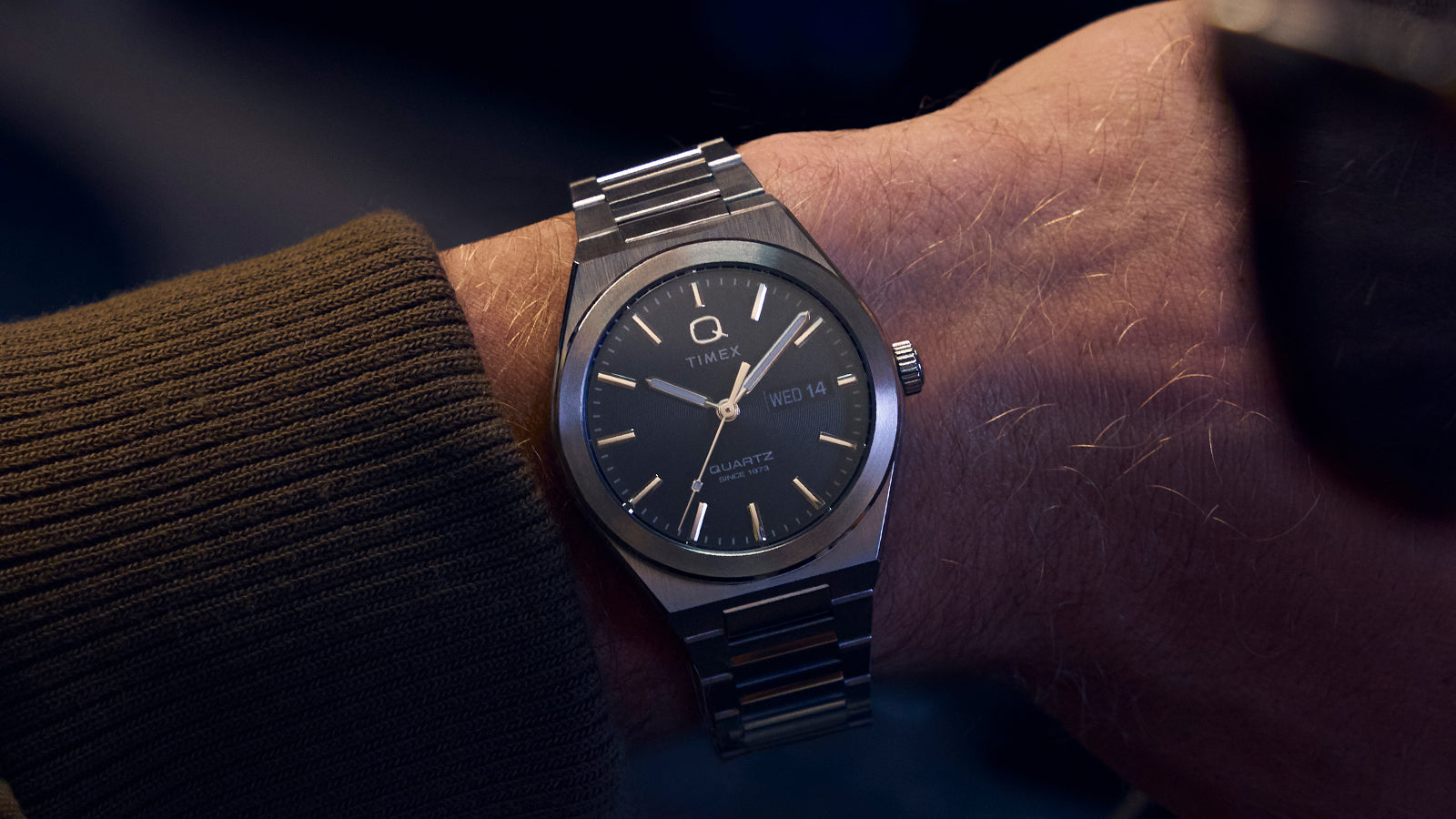 Wrist shot of the Q Timex® Continental Day and Date worn under a brown jacket sleeve. The stainless-steel bracelet and dark dial pair effortlessly for a timeless, understated style suited for both casual and formal wear.