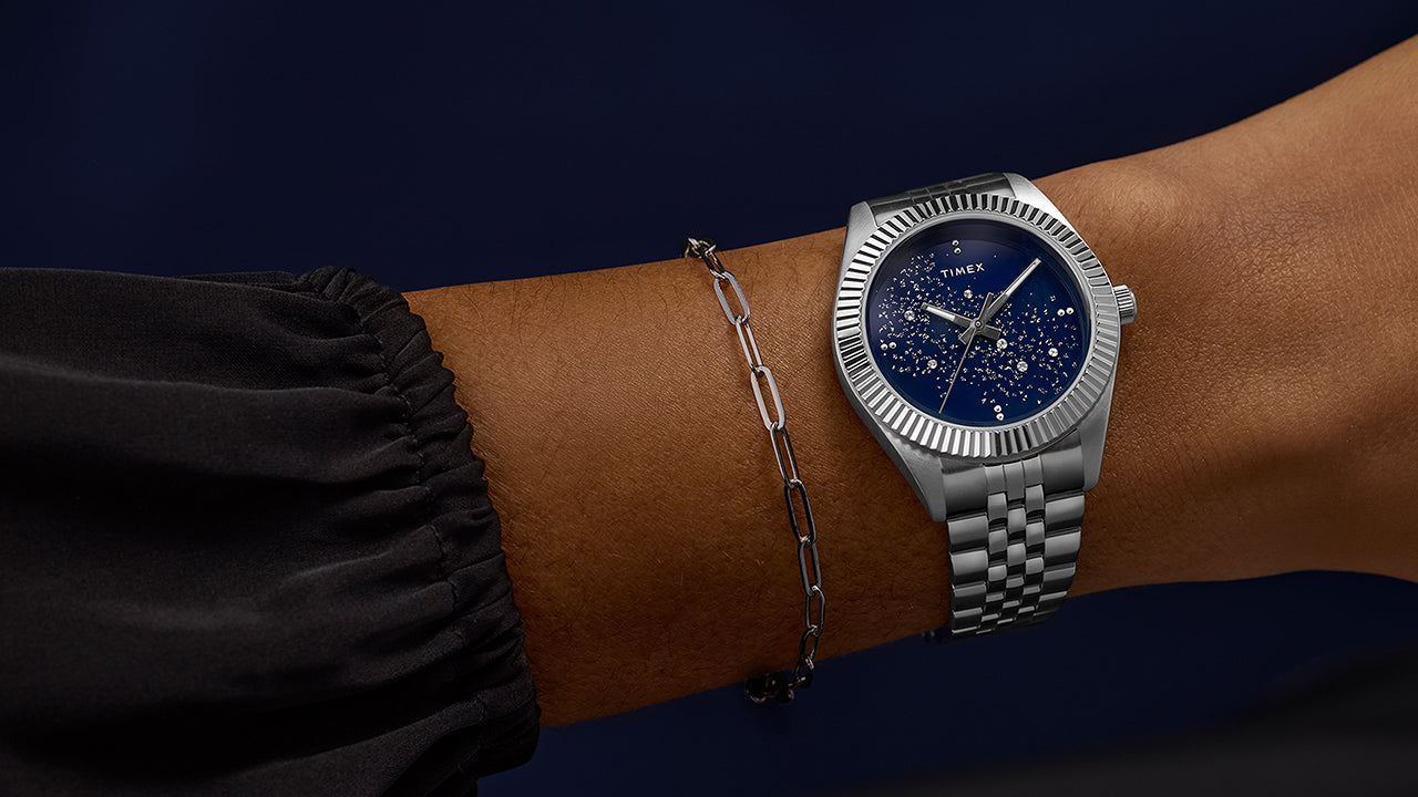 Timex Legacy watch worn on a wrist, featuring a blue starry dial and silver bracelet paired with a minimalist chain bracelet for an elegant look.