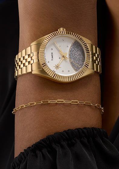 Timex Legacy gold-tone watch styled on wrist with a black sleeve and gold chain bracelet, showcasing its elegant crystal-accented dial and textured glitter detail.