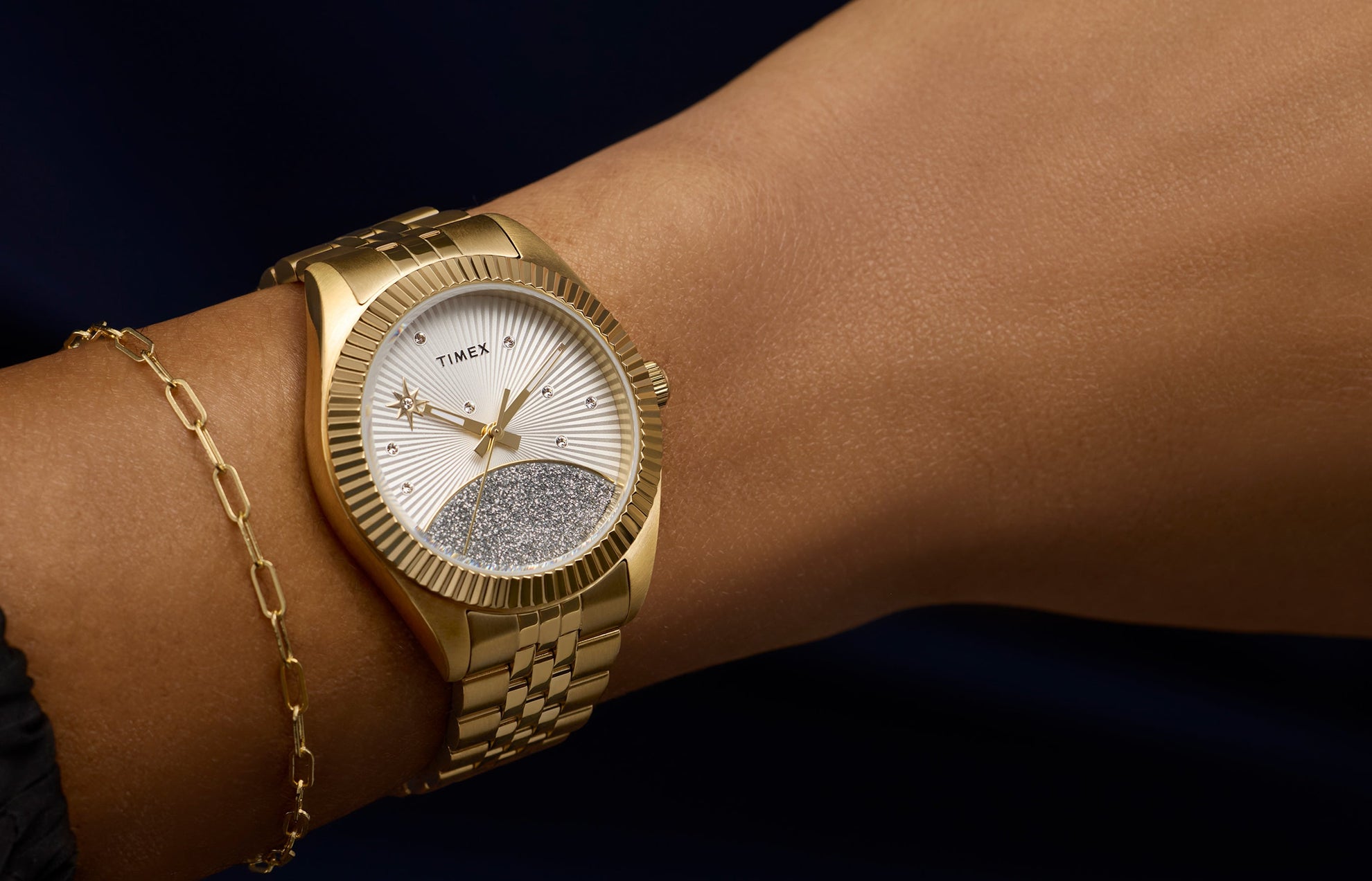 Timex Legacy watch in gold-tone stainless steel with a silver sunray dial featuring crystal accents and a glitter-textured lower half, worn on wrist with a delicate gold bracelet.