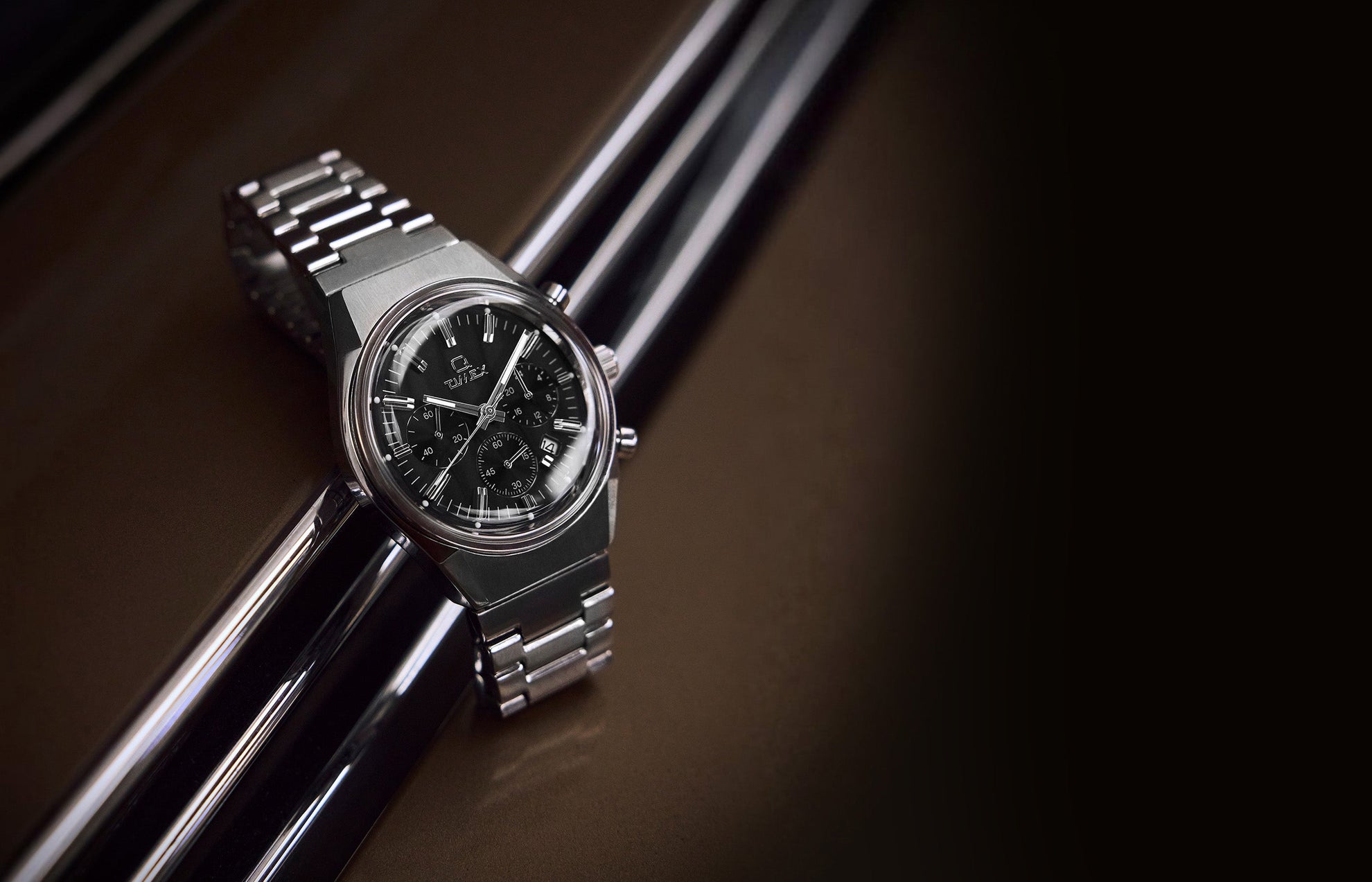 Q Timex Falcon Eye Chronograph watch with black dial, stainless-steel case, and bracelet, displayed on a glossy brown surface with chrome reflections.