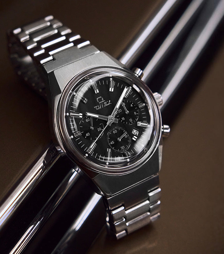 Q Timex Falcon Eye Chronograph watch with black dial, stainless-steel case, and bracelet, displayed on a glossy brown surface with chrome reflections.