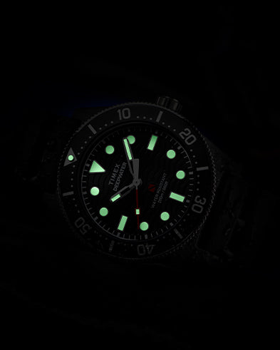Timex Deepwater dive watch glowing in the dark, showing bright green luminescent hour markers and hands against the black dial, with the red seconds hand and bezel numerals faintly visible.