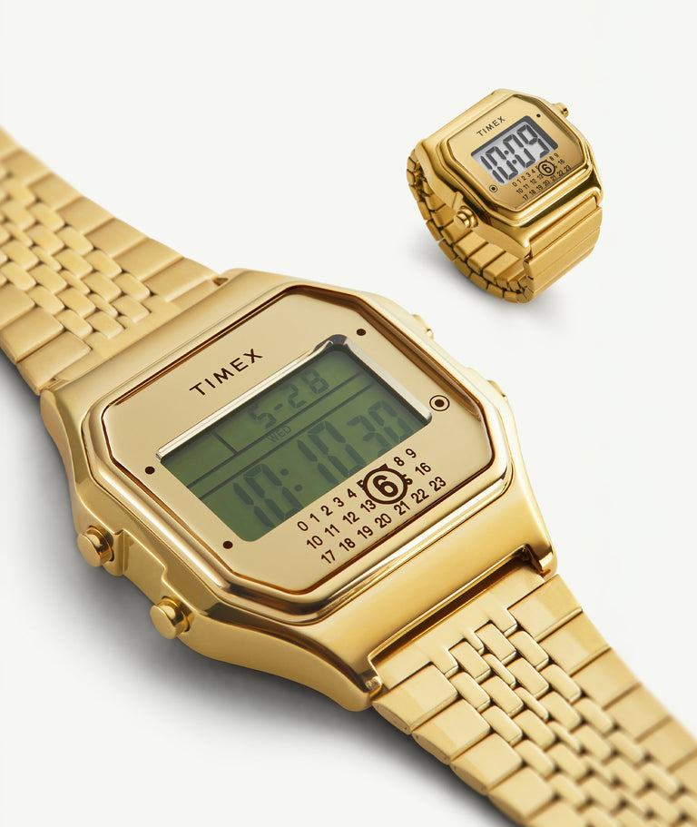 Two gold-tone Timex x MM6 digital watches displayed on a light background. The larger watch is shown close-up, highlighting its rectangular case, digital display with time, date, and calendar, and a gold metal bracelet. A smaller view of the same watch appears in the upper right, showing the full shape and stretch-style band.