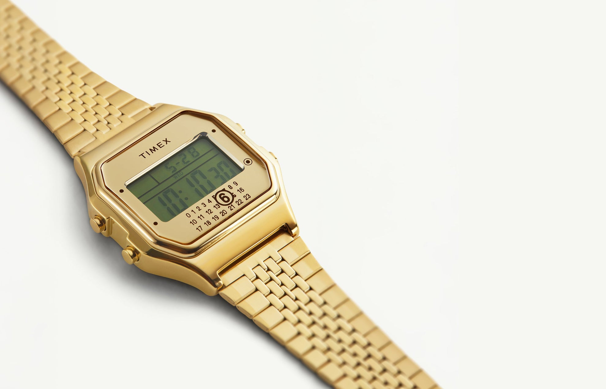 Close-up of the Timex x MM6 T80 digital wristwatch in gold-tone stainless steel, featuring a rectangular case, digital display showing the time 10:10:30, and signature MM6 calendar motif with the number 6 circled at the bottom of the dial.