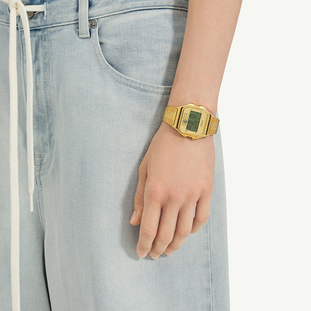The gold-tone Timex x MM6 T80 watch worn on a wrist, paired with light blue denim jeans, showing its retro digital aesthetic and comfortable bracelet fit.