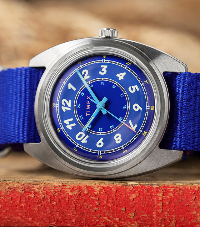 A Timex x Worn & Wound Whirlpool watch with a bright blue dial and matching blue fabric strap resting on a distressed red wooden surface, with thick coiled rope blurred in the background.