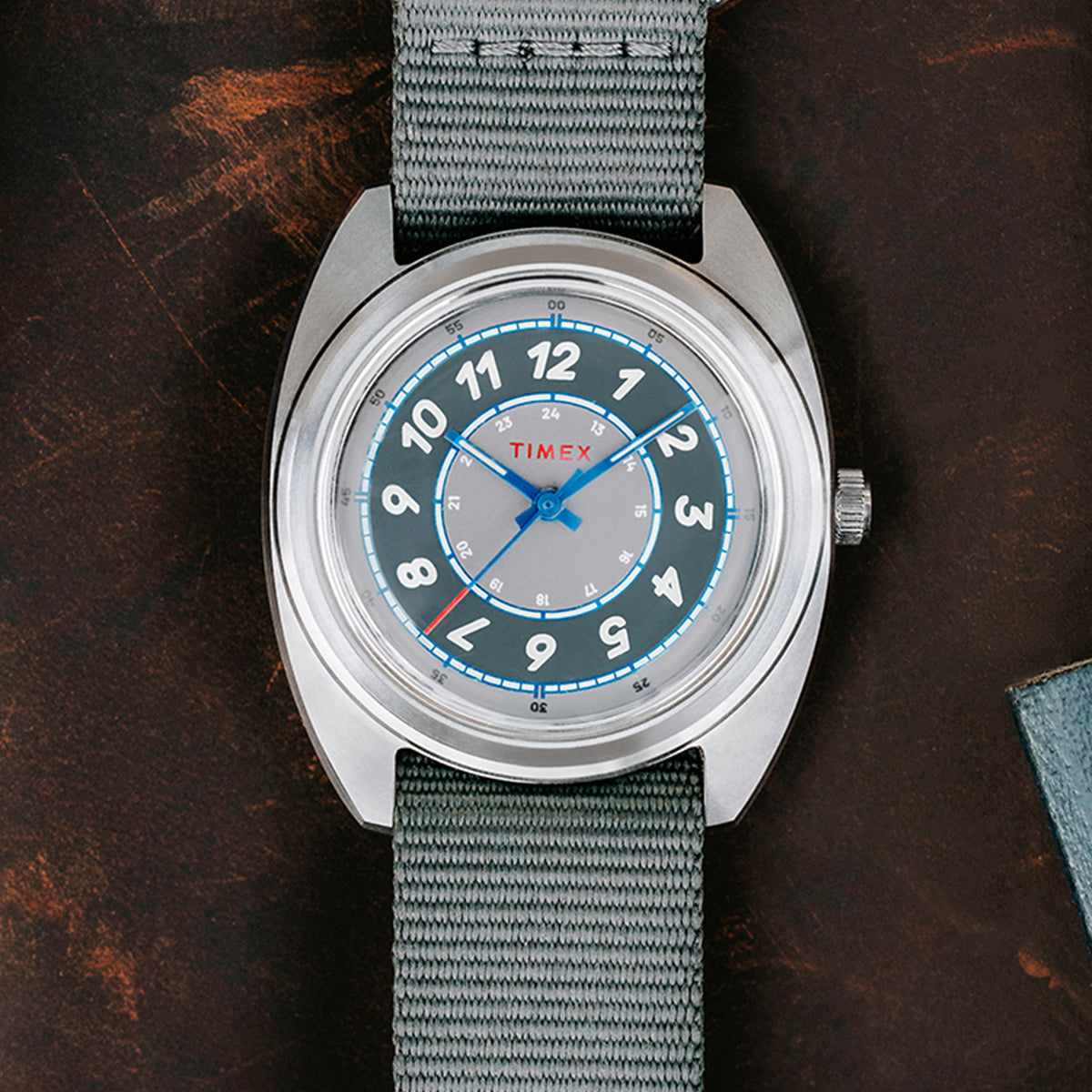 A top-down close-up of the Timex x Worn & Wound Storm watch showcasing its gray outer ring, silver center dial, bold cream numerals, and blue accents on a gray fabric strap.