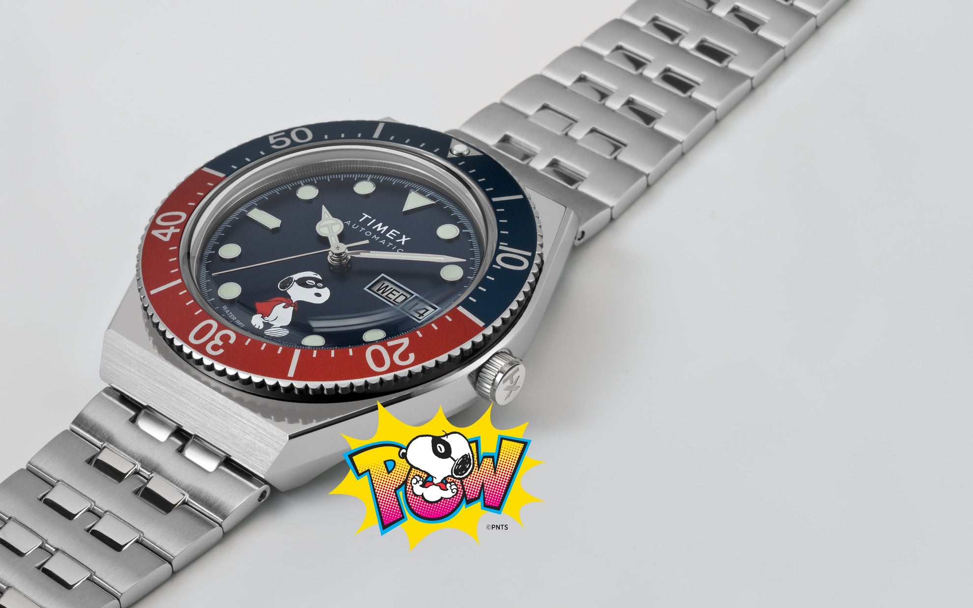 A Timex M79 Automatic x Peanuts watch with a blue dial featuring Snoopy dressed as a superhero. The watch has a stainless-steel bracelet, red and blue rotating bezel, and a “POW!” comic-style Snoopy illustration beneath it, blending playful design with classic dive watch styling.