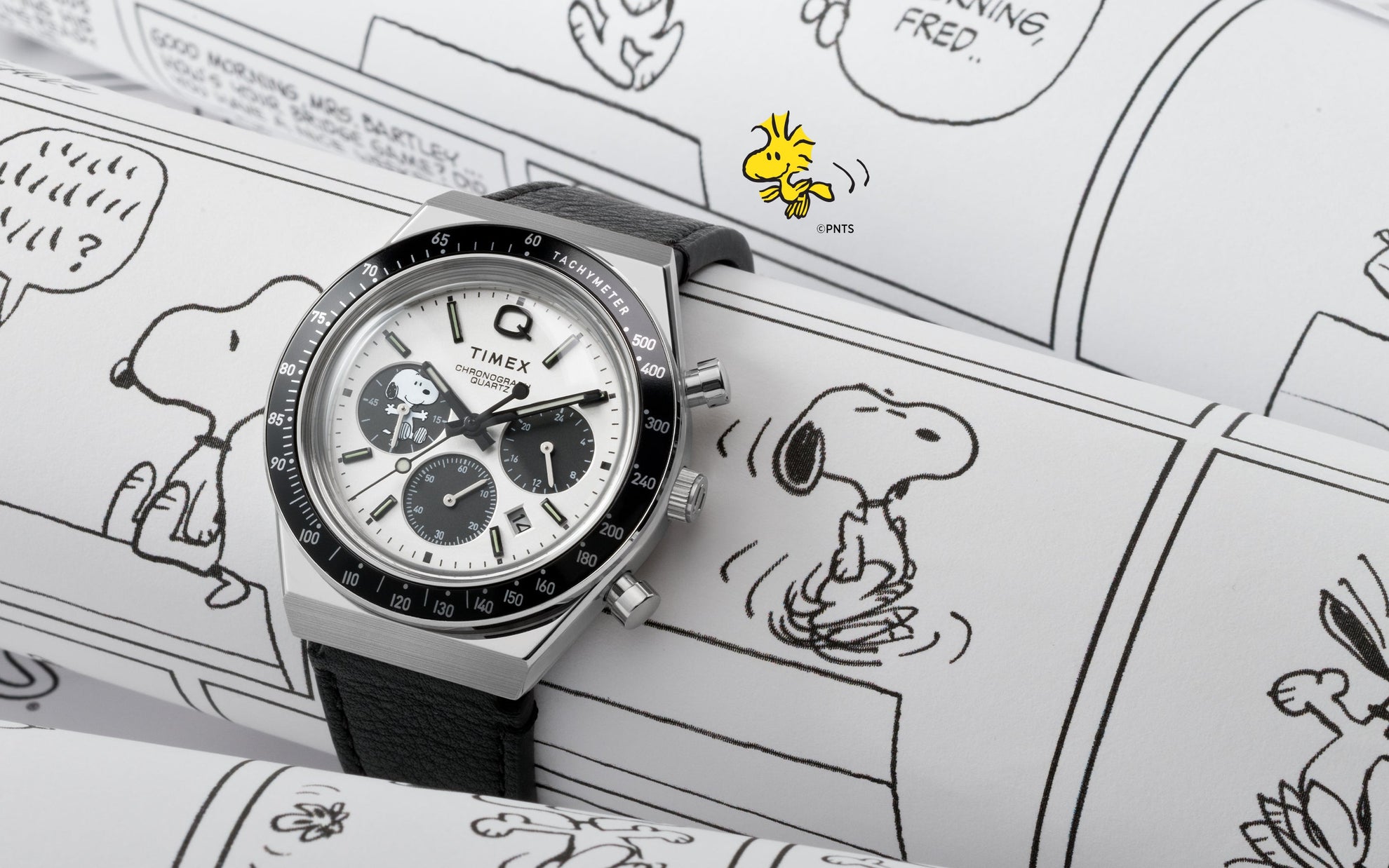 A Q Timex Chronograph x Peanuts Snoopy watch with a white dial, black sub-dials, and a black tachymeter bezel resting on Peanuts comic strips. The dial features Snoopy in one of the chronograph sub-dials, blending playful illustration with vintage racing-inspired design.