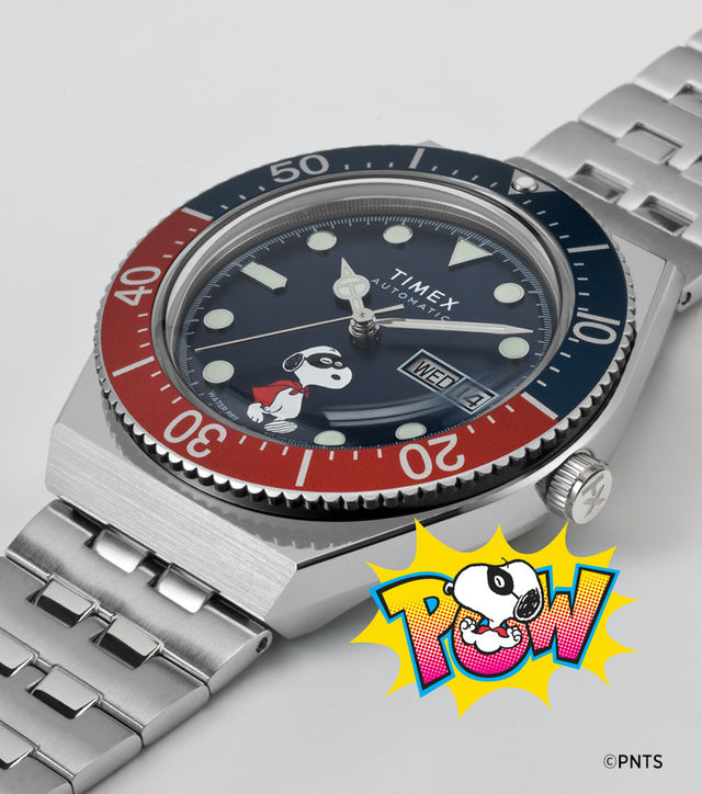 A Timex M79 Automatic x Peanuts watch with a blue dial featuring Snoopy dressed as a superhero. The watch has a stainless-steel bracelet, red and blue rotating bezel, and a “POW!” comic-style Snoopy illustration beneath it, blending playful design with classic dive watch styling.