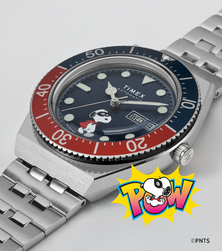 A Timex M79 Automatic x Peanuts watch with a blue dial featuring Snoopy dressed as a superhero. The watch has a stainless-steel bracelet, red and blue rotating bezel, and a “POW!” comic-style Snoopy illustration beneath it, blending playful design with classic dive watch styling.