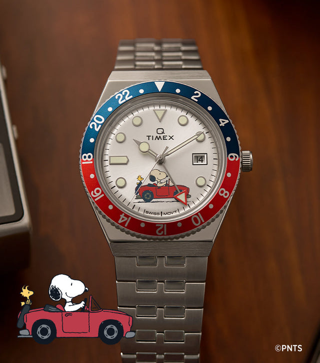 A Timex x Peanuts® Q Timex® GMT watch with a silver-tone stainless-steel bracelet and a red-and-blue 24-hour bezel displayed on a wooden desk. The silver dial features Snoopy driving a red convertible with Woodstock in the passenger seat, adding a whimsical touch to the retro GMT design.