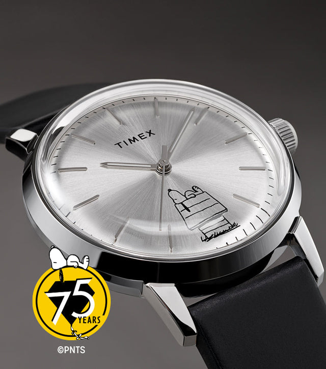 A Timex x Peanuts® Marlin® Automatic 75th Anniversary Edition watch with a silver sunburst dial and black leather strap displayed on a gray background. The dial features Snoopy lounging atop his doghouse in minimalist black line art, paired with the Peanuts 75th anniversary logo.