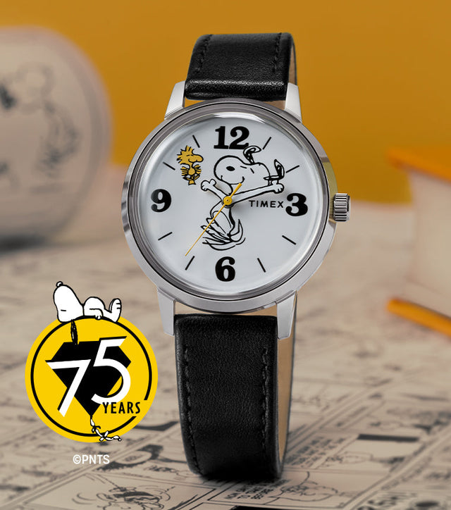 A Timex x Peanuts® Marlin® 75th Anniversary Edition watch with a white dial, black leather strap, and polished stainless-steel case displayed on a comic-themed background. The dial features Snoopy swinging a golf club with Woodstock flying nearby, and a yellow Peanuts 75 Years logo accent in the corner.