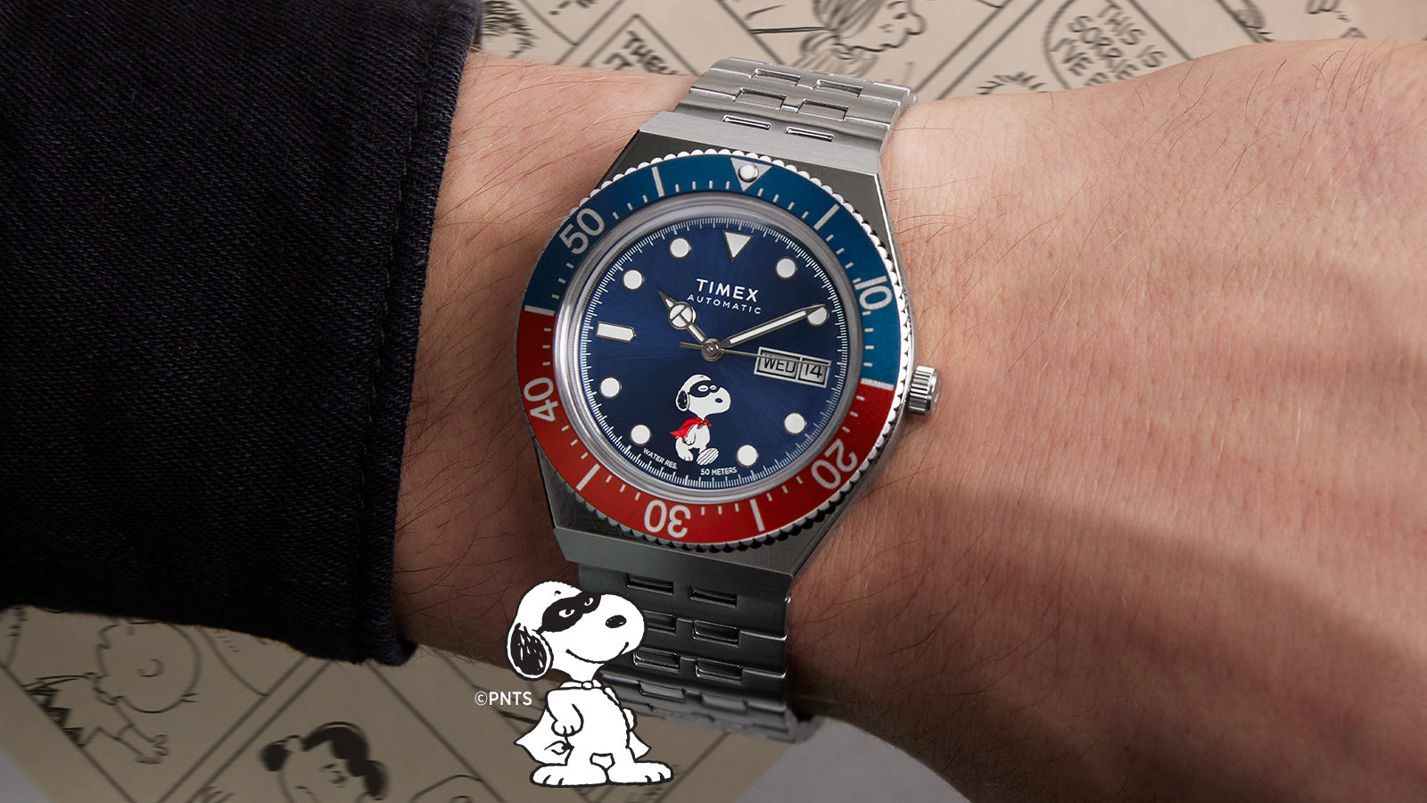 The Timex M79 Automatic x Peanuts watch shown on a wrist, highlighting its two-tone red and blue bezel, blue dial, and Snoopy superhero illustration at 6 o’clock. The playful design stands out against a Peanuts comic strip background.