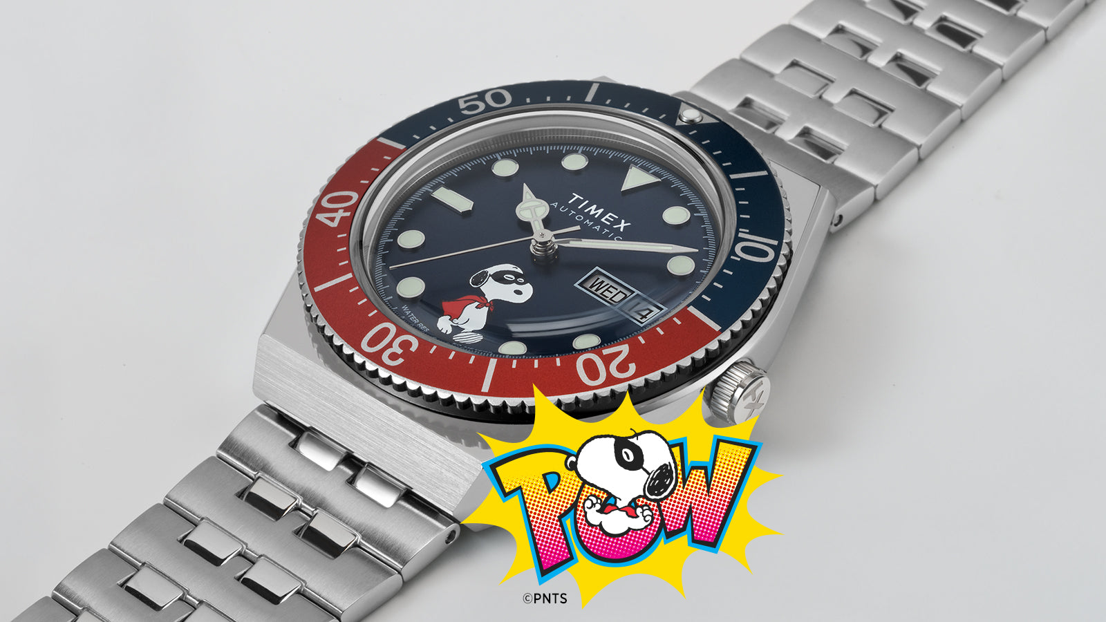 A Timex M79 Automatic x Peanuts watch with a blue dial featuring Snoopy dressed as a superhero. The watch has a stainless-steel bracelet, red and blue rotating bezel, and a “POW!” comic-style Snoopy illustration beneath it, blending playful design with classic dive watch styling.