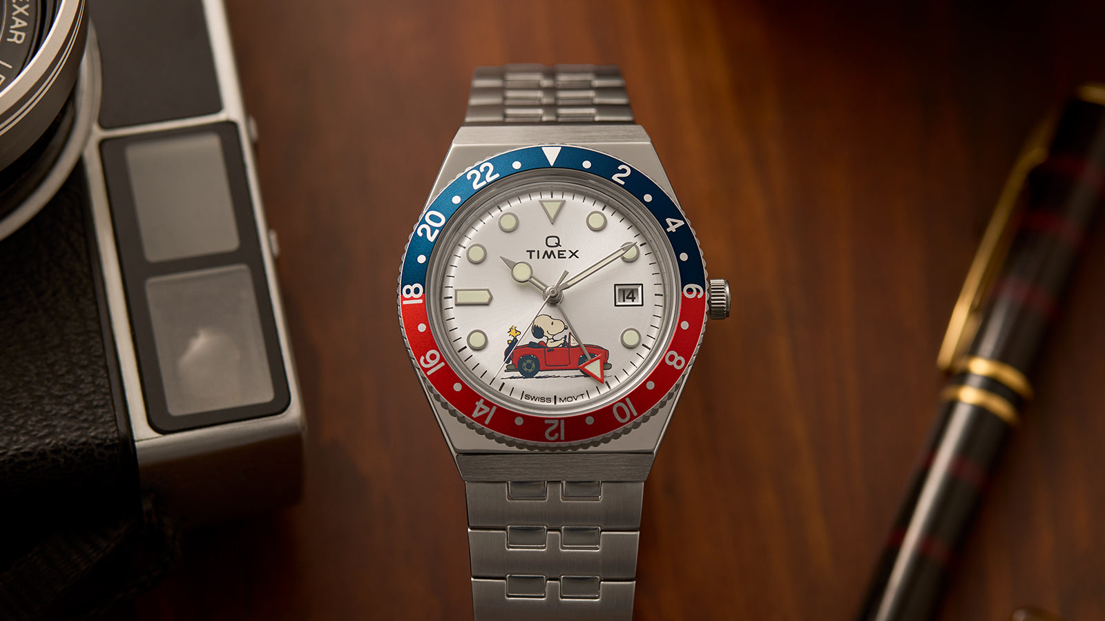 A Timex x Peanuts® Q Timex® GMT watch with a silver-tone stainless-steel bracelet and a red-and-blue 24-hour bezel displayed on a wooden desk. The silver dial features Snoopy driving a red convertible with Woodstock in the passenger seat, adding a whimsical touch to the retro GMT design.