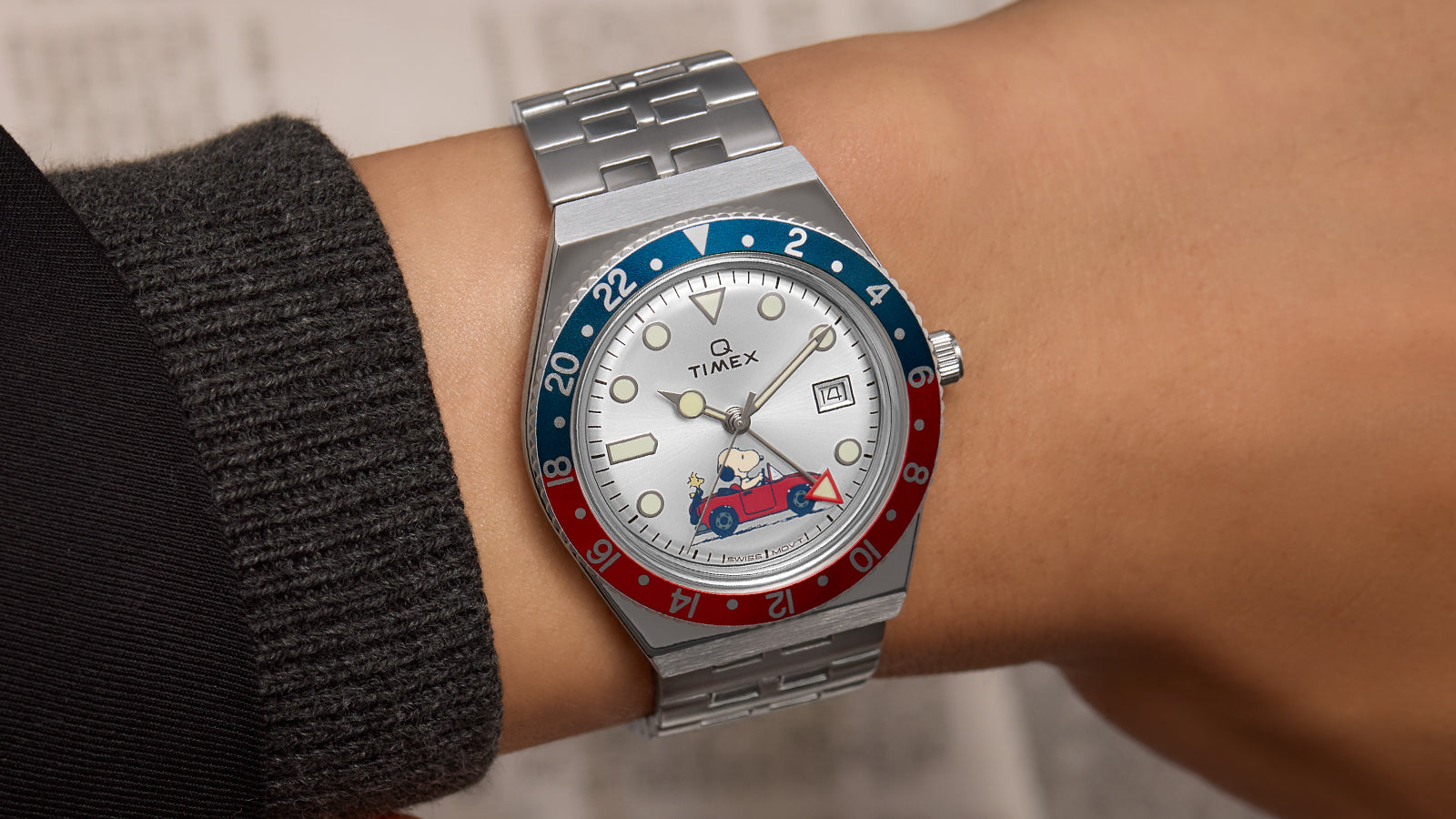 The Timex x Peanuts® Q Timex® GMT watch worn on a wrist, showing its brushed stainless-steel bracelet and silver dial. Snoopy and Woodstock are depicted driving a red car, with the two-tone red and blue GMT bezel and date window clearly visible.