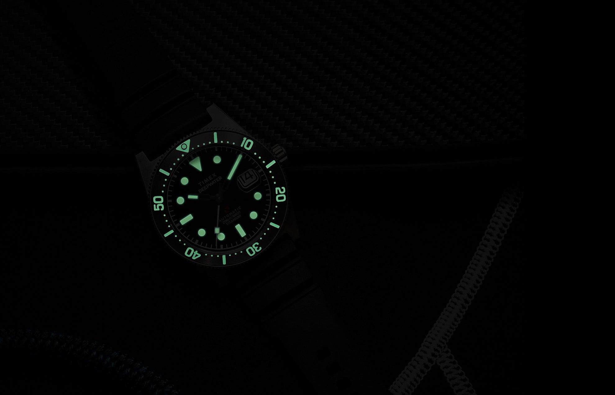 Timex Deepwater Reef 200 Titanium Automatic watch glowing in the dark, showing strong luminescence on the dial and bezel markers against a dark background.