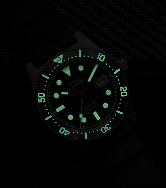 Timex Deepwater Reef 200 Titanium Automatic watch glowing in the dark, showing strong luminescence on the dial and bezel markers against a dark background.