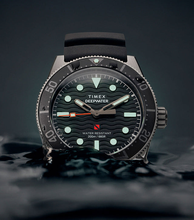 Timex Deepwater Reef 200 dive watch emerging from dark water, featuring a black dial with luminous markers and hands, black rubber strap, and 200-meter water resistance rating.