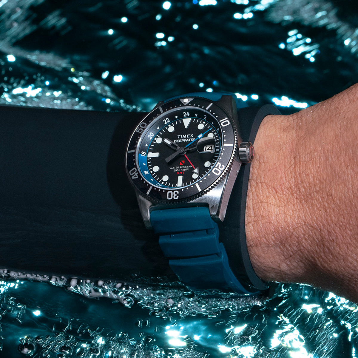 Diver wearing the Timex Deepwater Reef 200 watch underwater, the blue strap and stainless steel case reflecting light as bubbles rise around them.