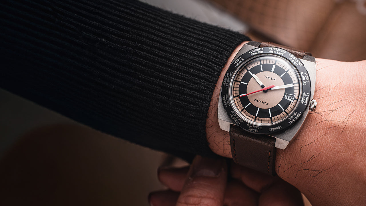 Lifestyle image of the Q Timex® 1972 World Time watch on wrist, displaying the bold tan-and-black world time dial, brushed steel case, red seconds hand, and brown leather strap paired with a dark sweater.