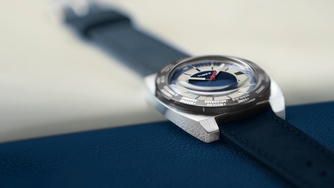 Angled view of the Q Timex® 1972 World Time watch featuring a navy blue leather strap, brushed stainless-steel case, and cream-and-blue dial with a red seconds hand.