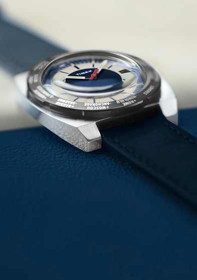 Angled view of the Q Timex® 1972 World Time watch featuring a navy blue leather strap, brushed stainless-steel case, and cream-and-blue dial with a red seconds hand.