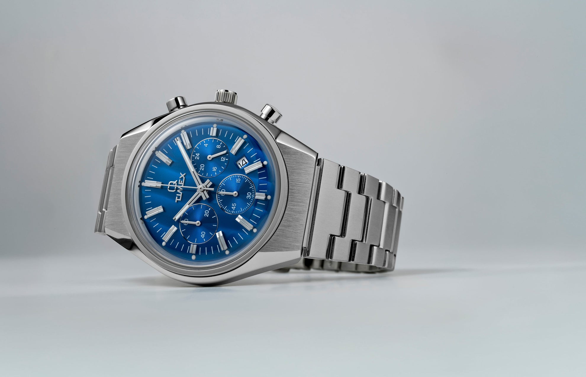 Q Timex Falcon Eye Chronograph with a brushed stainless-steel case and bracelet, featuring a metallic blue dial with three subdials and a date window. The watch is displayed on a light gray background.