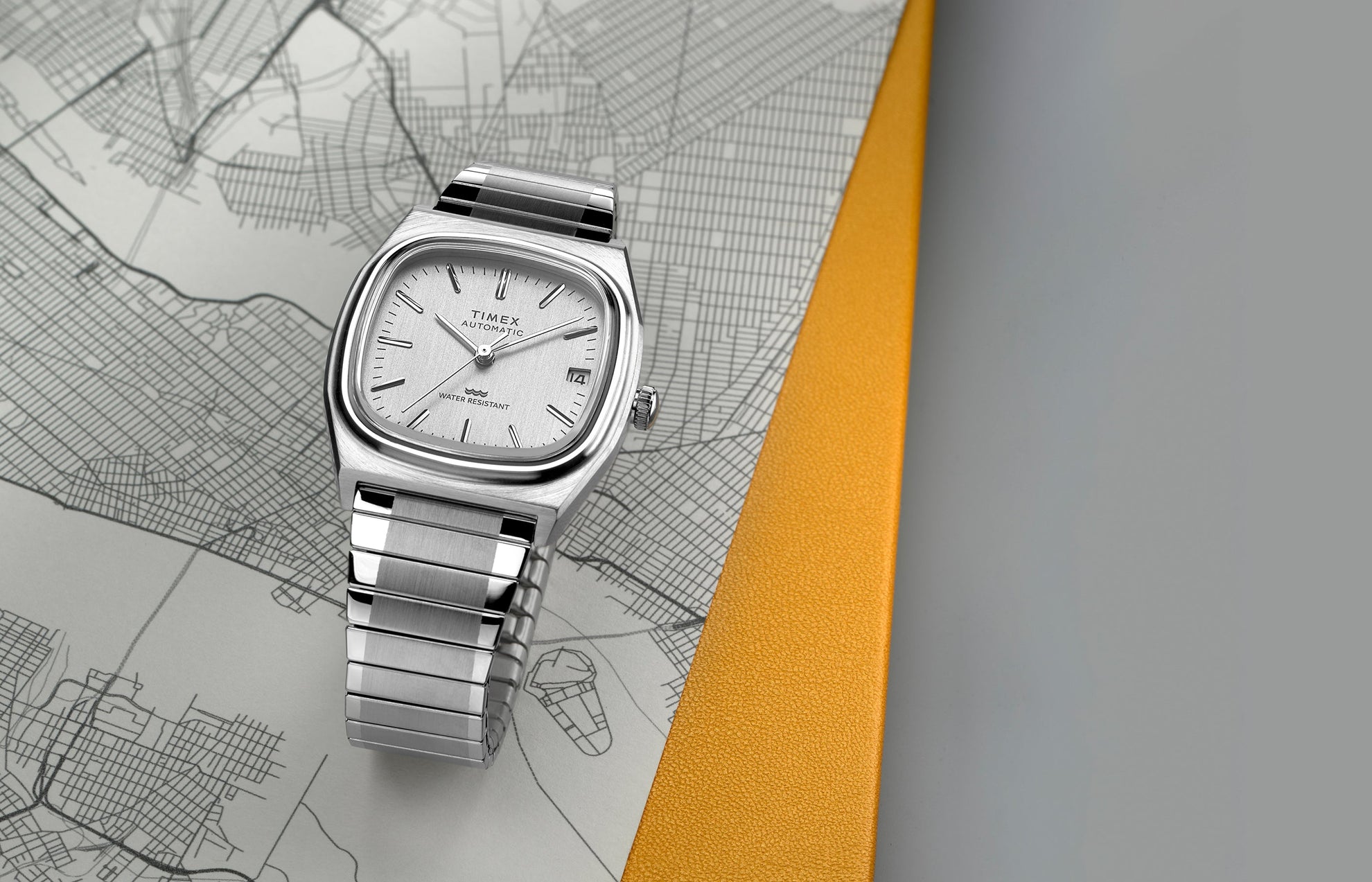 Timex® Automatic 1983 E Line Reissue watch with a brushed stainless-steel case and integrated bracelet, featuring a silver dial with minimalist hour markers and date window at 3 o’clock. The watch is displayed on a city map background with a yellow accent.