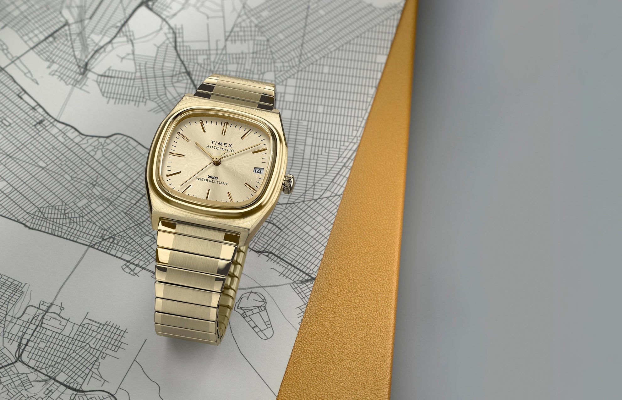 Timex® Automatic 1983 E Line Reissue watch in gold-tone stainless steel with an integrated bracelet and champagne dial. The watch is displayed on a detailed city map with a yellow textured accent in the background.