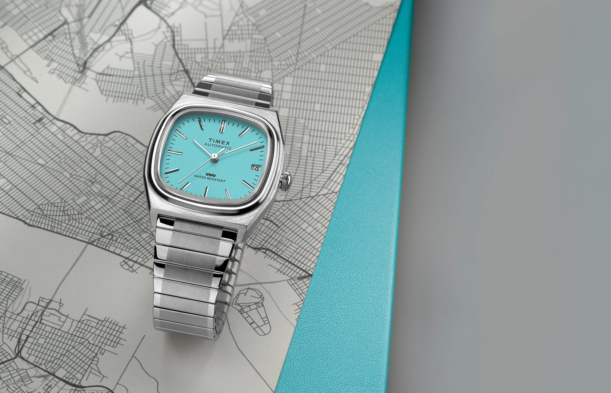 Timex® Automatic 1983 E Line Reissue watch with a stainless-steel case and bracelet, featuring a bright turquoise dial with silver-tone hour markers and hands. The watch is displayed on a map background with a teal accent.