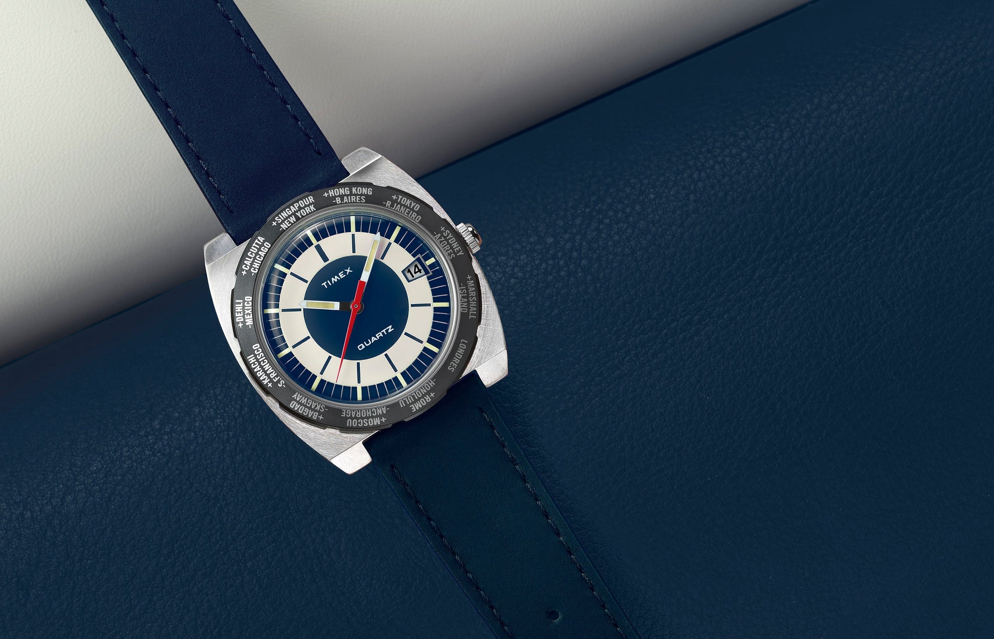 Top-down view of the Q Timex® 1972 World Time watch with navy blue strap and blue-and-cream dial resting on blue textured leather.