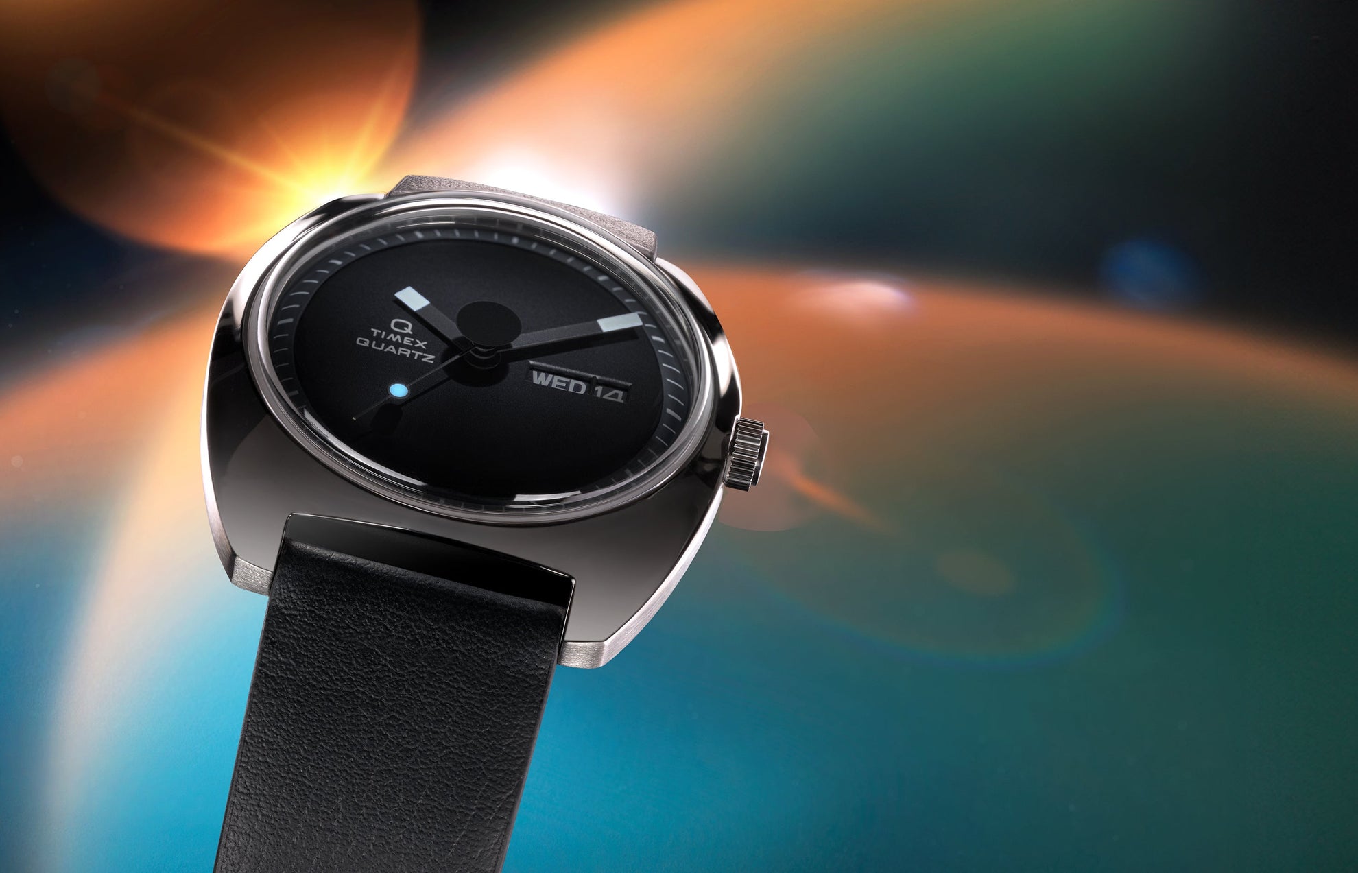 Angled view of the Q Timex® 1975 Enigma watch with a black dial, polished stainless-steel case, and black leather strap set against a glowing space-inspired background.