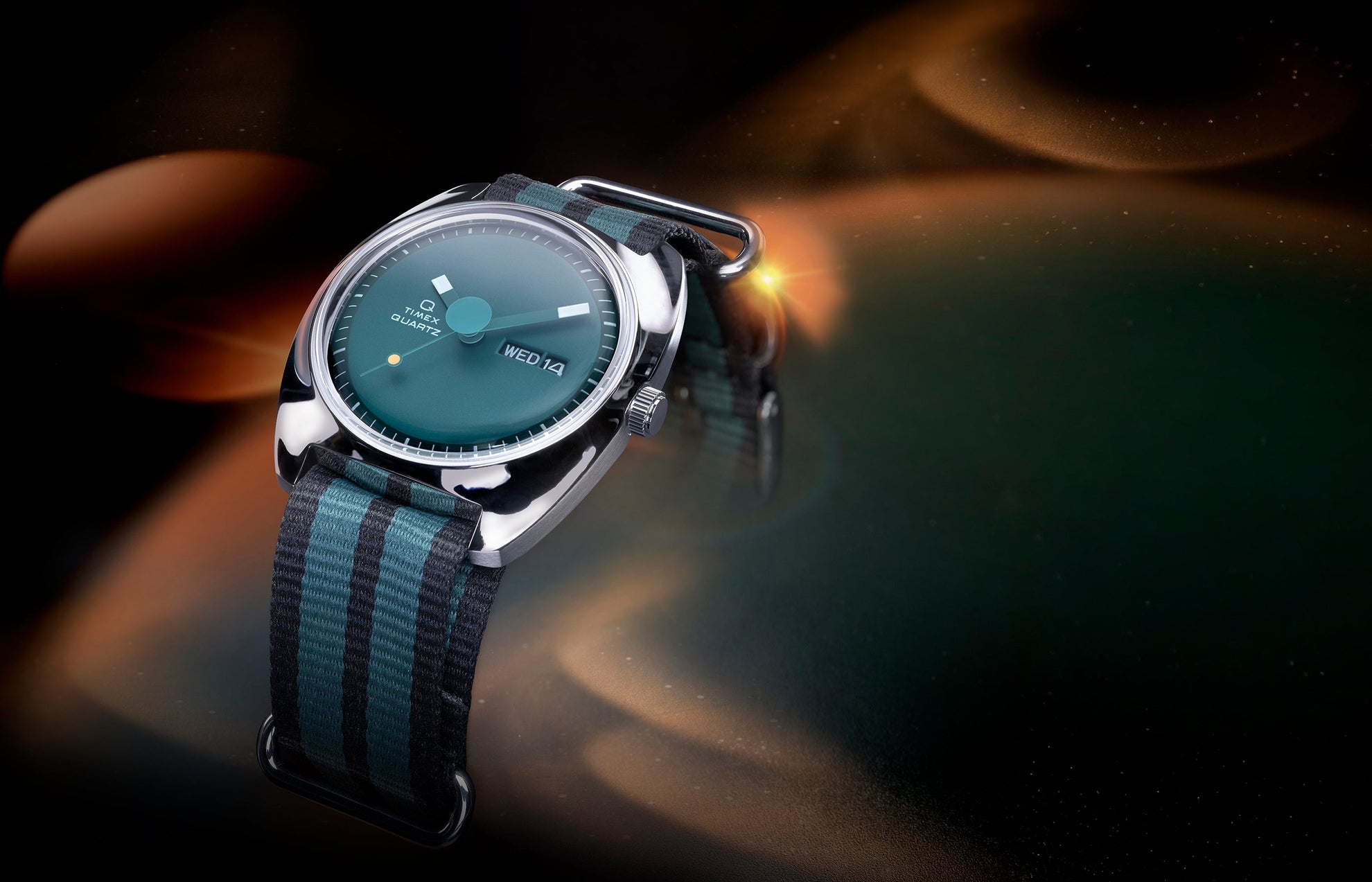 Angled view of the Q Timex® 1975 Enigma watch with a teal dial, polished silver-tone case, and black-and-green striped fabric strap, set against a glowing cosmic-inspired background.