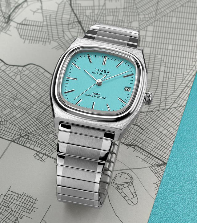 Timex® Automatic 1983 E Line Reissue watch with a stainless-steel case and bracelet, featuring a bright turquoise dial with silver-tone hour markers and hands. The watch is displayed on a map background with a teal accent.