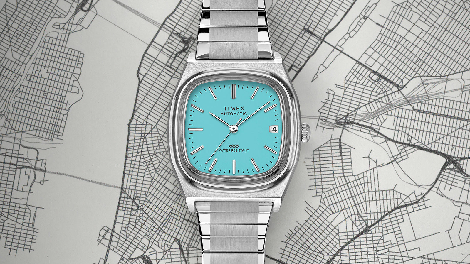 Top-down view of the Timex® Automatic 1983 E Line Reissue showcasing its turquoise dial, cushion-shaped case, and brushed stainless-steel bracelet positioned over a city map.