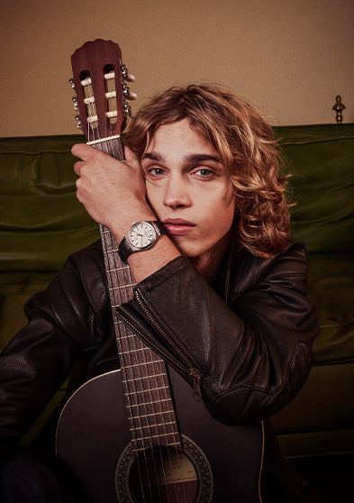 Model seated on a green sofa, embracing an acoustic guitar while wearing the Q Timex® Continental GMT watch, styled in a black leather jacket for a relaxed, vintage-inspired look.