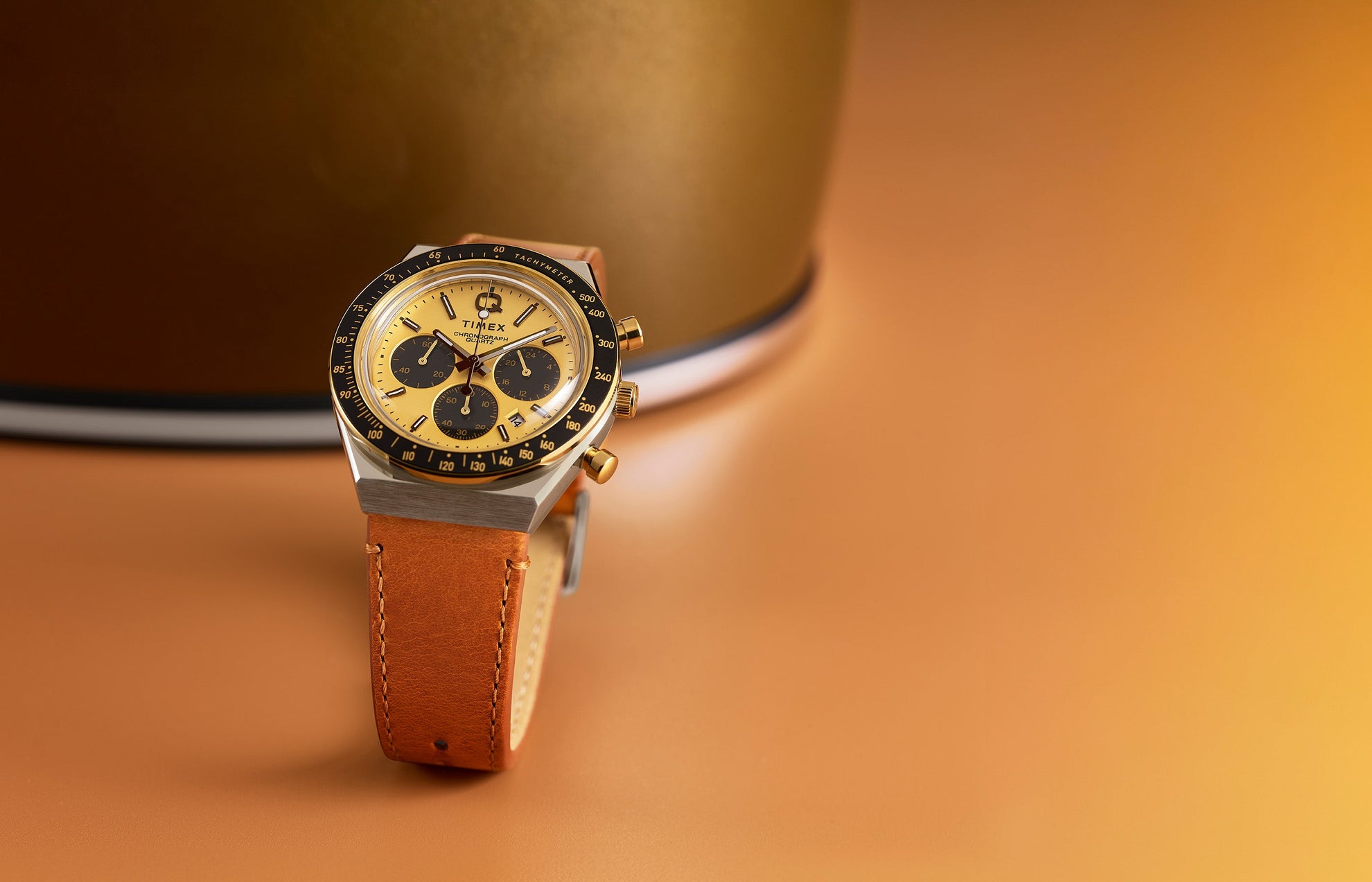 Q Timex Chronograph featuring a gold-tone dial, black sub-dials, and tan leather strap displayed upright on a warm gold background.