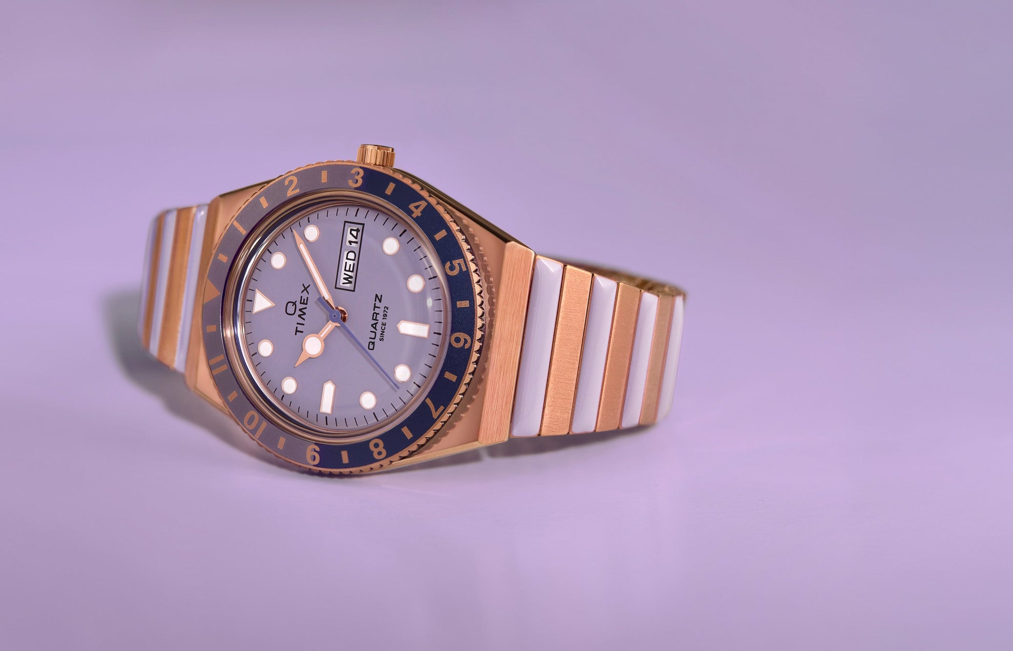 Q Timex® watch in rose gold-tone with a lilac dial and blue bezel, featuring a two-tone stainless steel bracelet with white center links, displayed against a soft lavender background.