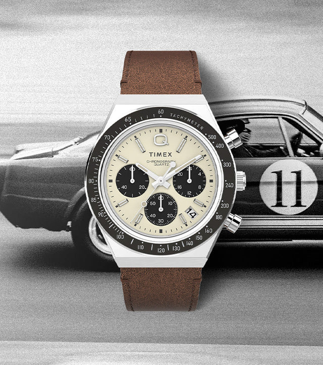 Q Timex Chronograph watch with a cream dial, black sub-dials, and brown leather strap displayed over a vintage racing car background, evoking a classic motorsport aesthetic.