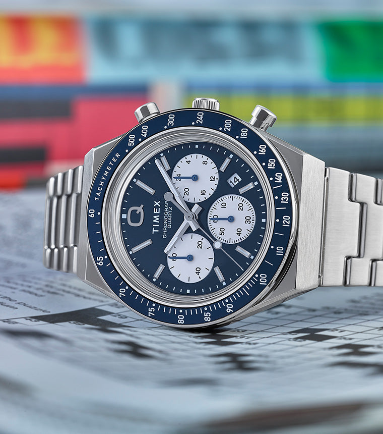 Q Timex Chronograph with a blue dial, white sub-dials, and stainless steel bracelet resting on a folded newspaper with colorful pages in the background.