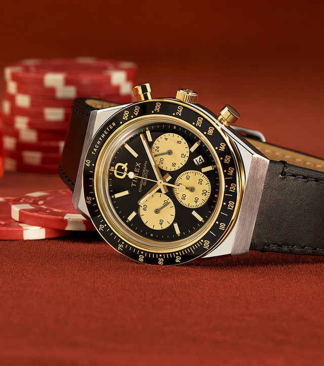 Q Timex Chronograph with a black dial and gold-tone sub-dials on a black leather strap, resting on a poker table with red chips and dice in the background.